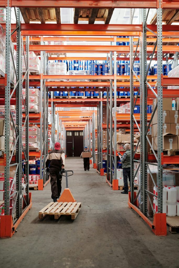 pexels photo 4483772 4483772 Workers arrange inventory on shelves in a large industrial warehouse.